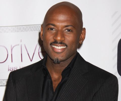 Romany Malco in a black suit smiles at the camera.